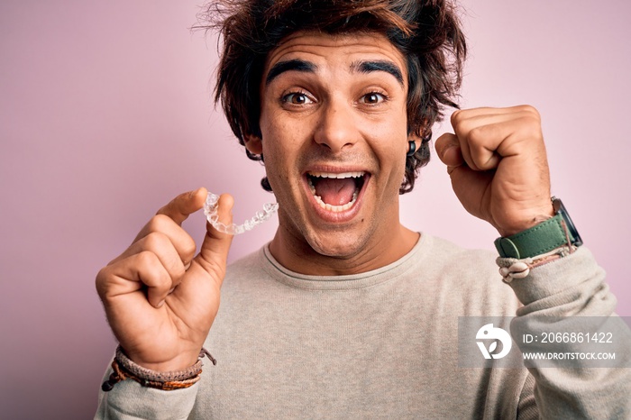 Young handsome man holding aligner standing over isolated pink background screaming proud and celebrating victory and success very excited, cheering emotion