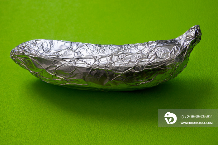 Banana covered with aluminum foil on a green background, irony of degradable bio packaging, nature manipulated by man.