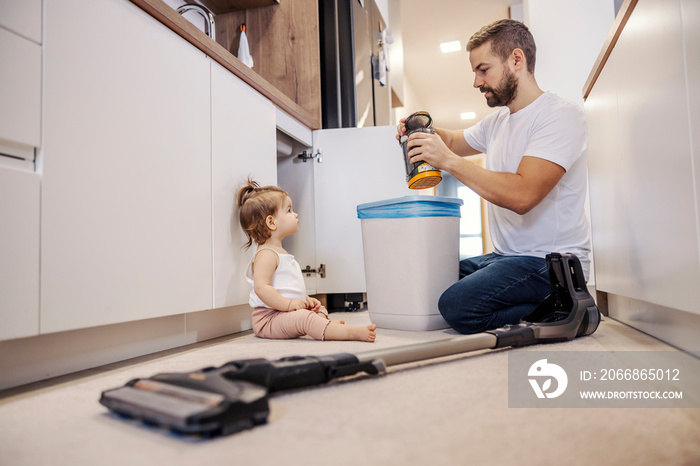 A man is cleaning dust from vacuum cleaner and putting dust into a garbage can while his baby girl is watching him.