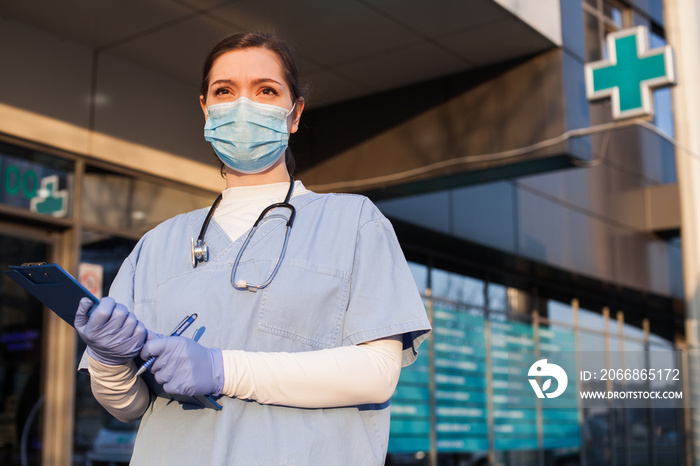 Young female doctor standing in front of healthcare facility, wearing protective face mask and PPE equipment, holding medical patient clipboard, COVID-19 pandemic crisis