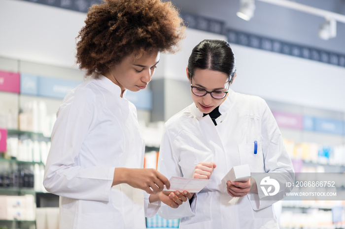 Two female pharmacists reading the information from the pack of a new product while comparing two different medicines regarding indications and side effects