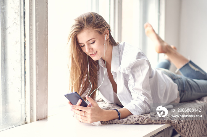 girl lies on the window and listens to music or podcast on her smartphone. digital detox concept