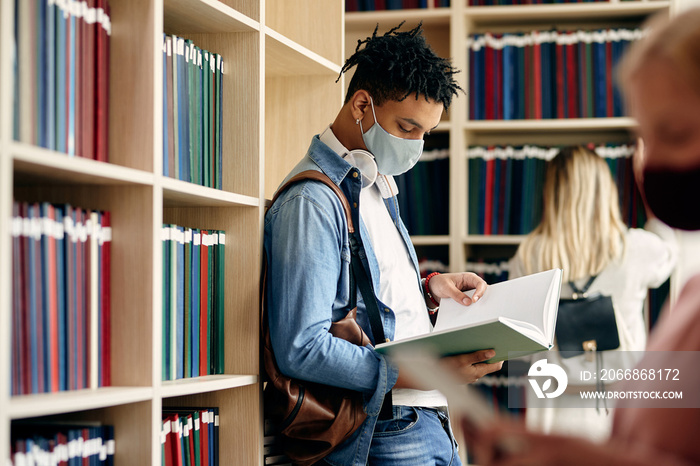 Black university student with face mask reading from a book while learning in library.