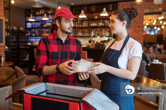 Young waitress in workwear passing plastic container with fresh food to courier