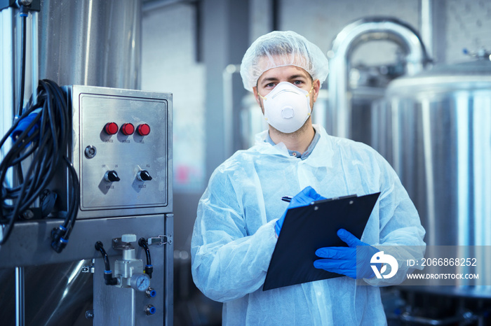 Factory worker in white protective uniform with hairnet and mask standing by industrial machine. Technologist checking production.