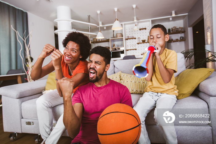 Happy African American family of three watching tv and cheering basketball games on sofa at home.