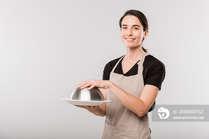 Happy young brunette waitress in apron keeping hand on cover of cloche