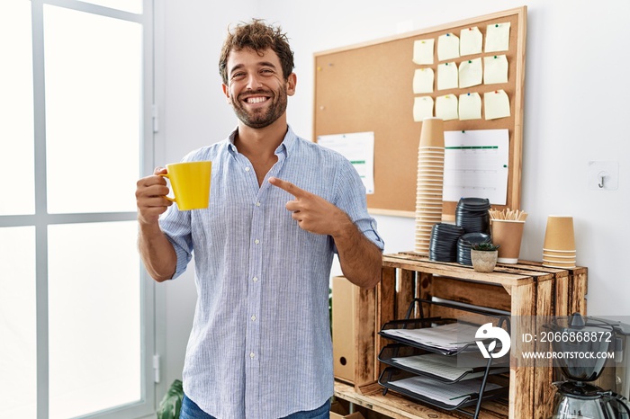 Young handsome man drinking a cup coffee at the office smiling happy pointing with hand and finger