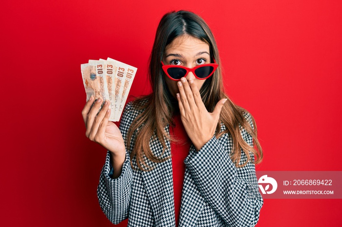 Young brunette woman holding united kingdom 10 pounds banknotes covering mouth with hand, shocked and afraid for mistake. surprised expression