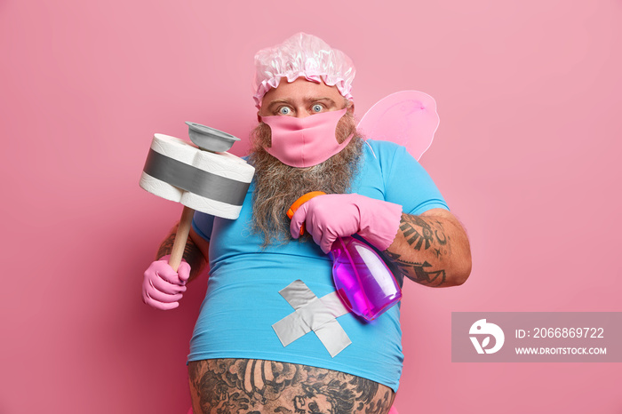 Scared bearded man helper ready to clean your house stares fearful at camera poses with detergent and plunger busy cleaning room poses against pink background. Funny fairy of purity. Housekeeping