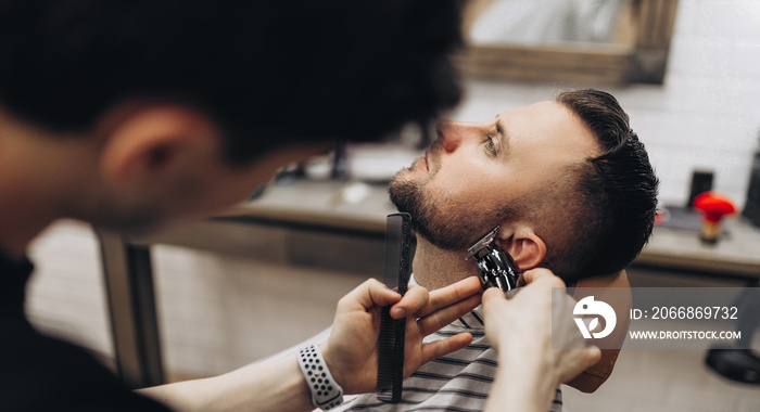 Hipster man sitting in armchair barber shop while hairdresser shaves beard with scissors.