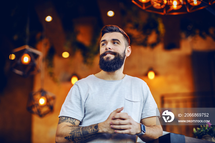 Low angle view of attractive smiling caucasian bearded tattooed hipster leaning on table in cafe and looking away.