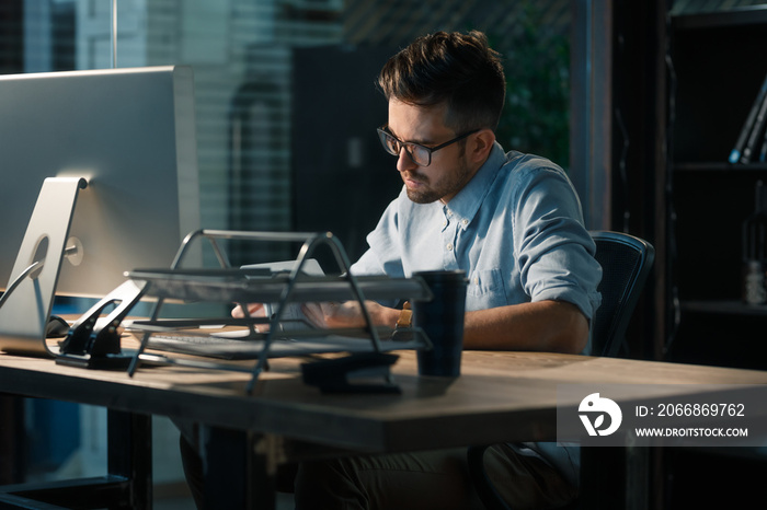 Young man in glasses working late alone at desktop in office taking notes and reading information.