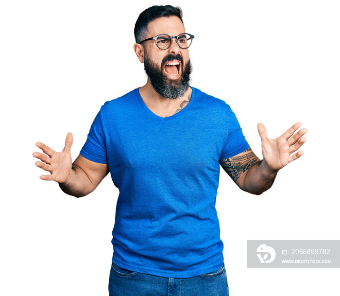 Hispanic man with beard wearing casual t shirt and glasses crazy and mad shouting and yelling with aggressive expression and arms raised. frustration concept.