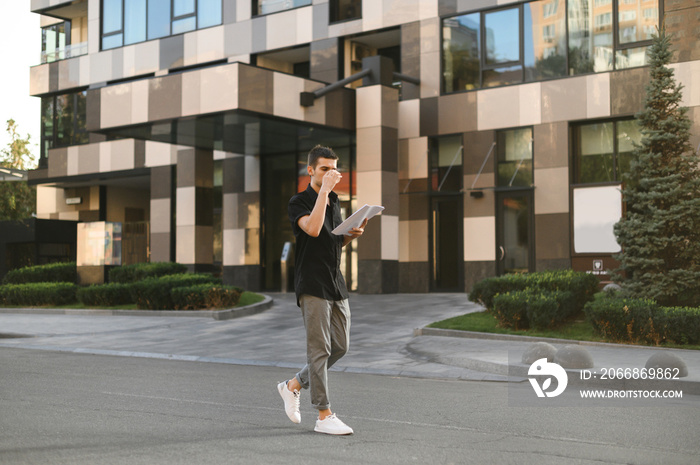 Portrait of a fashionable man in a black shirt and gray pants walking down the street with business papers in his hands and drinking coffee. Full length photo of stylish man walking down the street