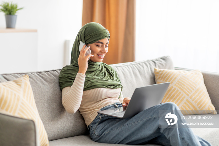 Cheerful young Arab woman in hijab using laptop for distance job, calling on smartphone, communicating online from home