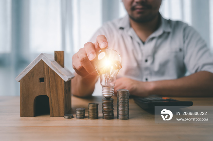 male hand holding a light bulb with coins stacking and house model on desk. The concept of installment and reduction of home loan interest, saving energy and money concept.