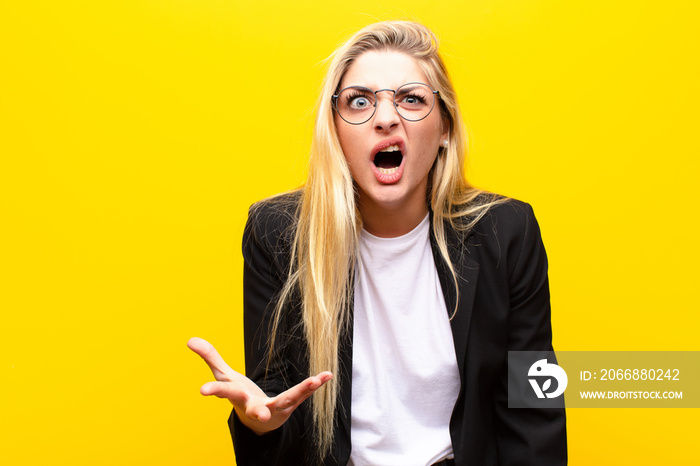 young pretty blonde woman looking angry, annoyed and frustrated screaming wtf or what‚Äôs wrong with you against yellow wall