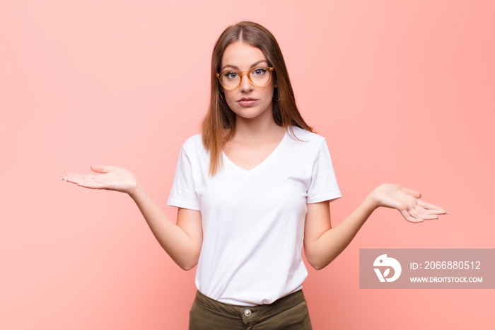 young red head woman feeling puzzled and confused, unsure about the correct answer or decision, trying to make a choice against flat wall