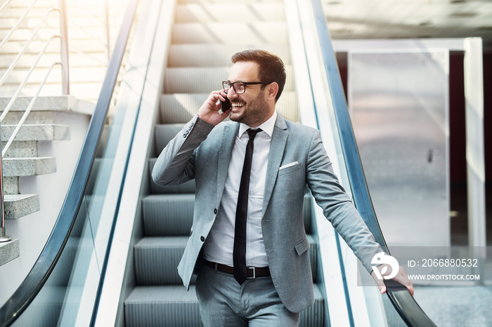 Young modern successful businessman talking on the telephone while standing on a escalator. Smiling about good business offer.