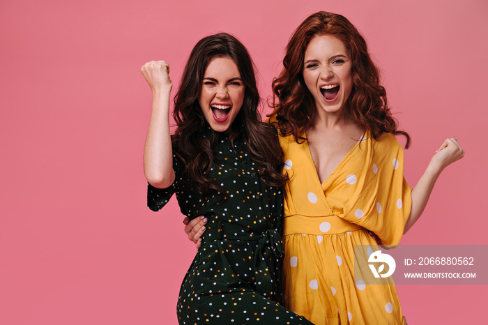 Optimistic women in bright dresses happily shout on pink background. Joyful girlfriends with curly hair and in polka dot dresses hugging