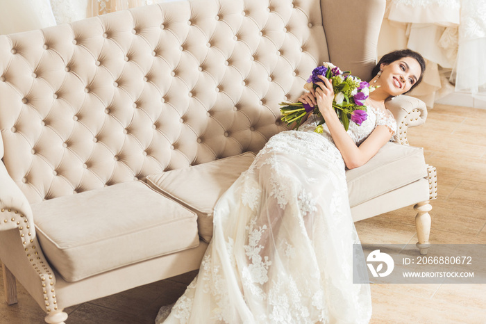 Young bride lying on sofa with flowers bouquet in wedding atelier