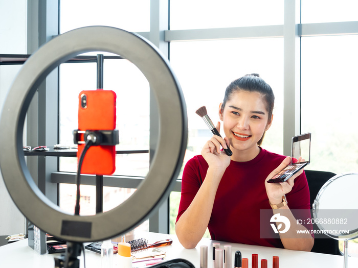 Asian beauty blogger woman use brush and multicolor eyeshadow for makeup and review product by mobile phone camera live on social media and record within artist cosmetic and skincare on desk in studio