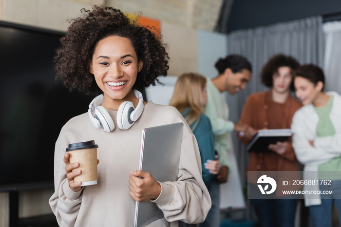 cheerful african american woman with disposable cup, headphones and laptop near blurred multiethnic classmates.