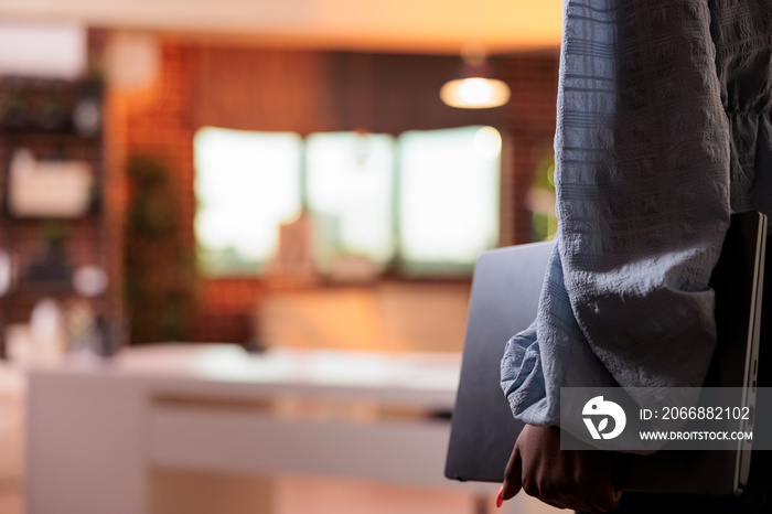 African american woman in casual clothes carrying laptop in home office with modern interior and beautiful warm sunset light. Female entrepreneur holding portable computer, side view