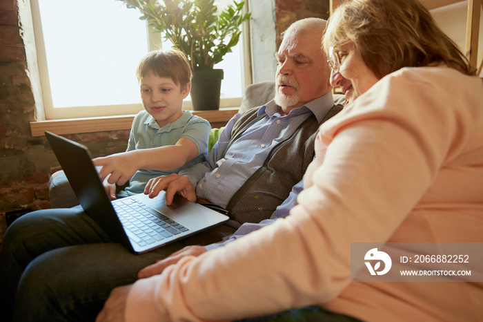 Life portrait of friendly family, grandparent and their grandson sitting on sofa and spending time together, using modern gadgets, talking, studying