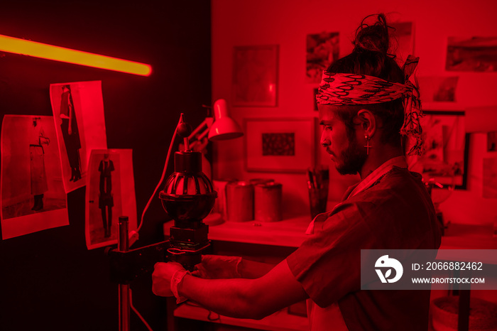 Young man working with film photography in red lit darkroom using photo enlarger to project image onto paper