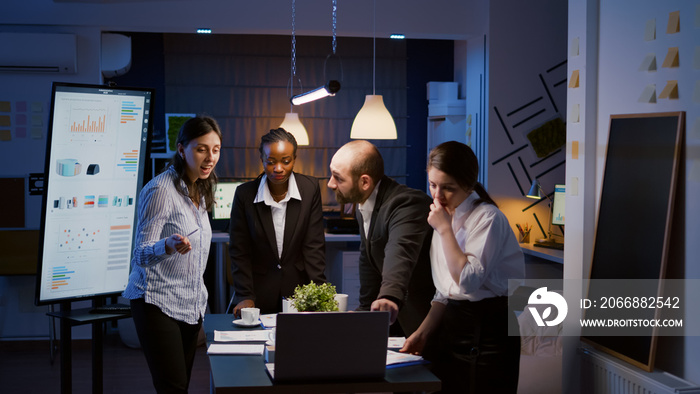 Multi ethnic businesspeople brainstorming company ideas checking graphs statistics using laptop standing at conference table. Diverse teamwork working at management presentation in meeting room