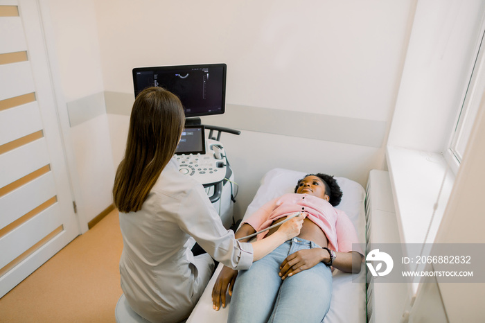 Young woman doctor looks at the ultrasound screen in a modern clinic, while performing ultrasound for young African pregnant woman