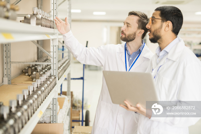 Two bearded technicians wearing lab coats standing at shelf of spacious manometer factory warehouse and taking inventory, one of them holding laptop in hands