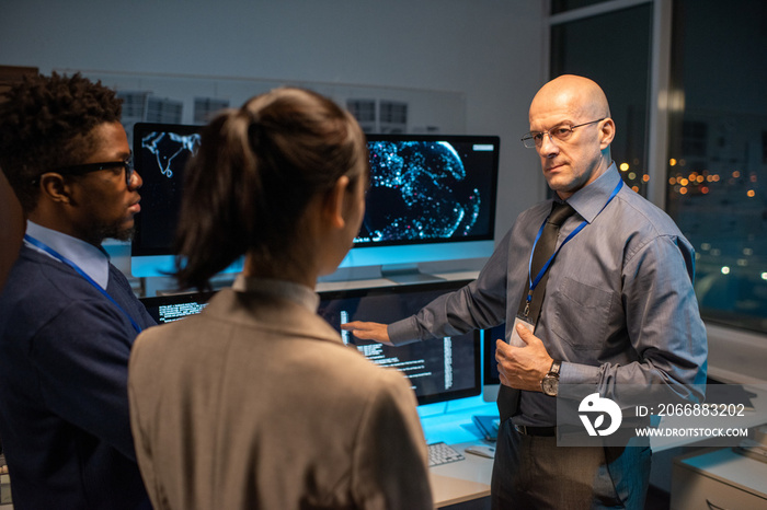 Bald mature director of software company giving instructions to young intercultural subordinates while pointing at data on computer screen