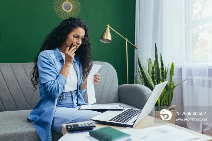 Serious and thoughtful businesswoman working at home with documents, hispanic woman in casual clothes using laptop at work sitting on sofa in living room with bills and contracts.