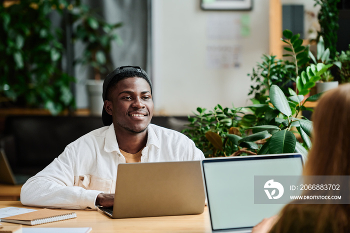 Young smiling African American businessman with laptop looking at colleague sitting in front of him during network and discussion