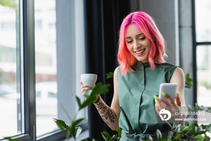 cheerful woman with pink hair and piercing holding cup of coffee while looking at smartphone in office.