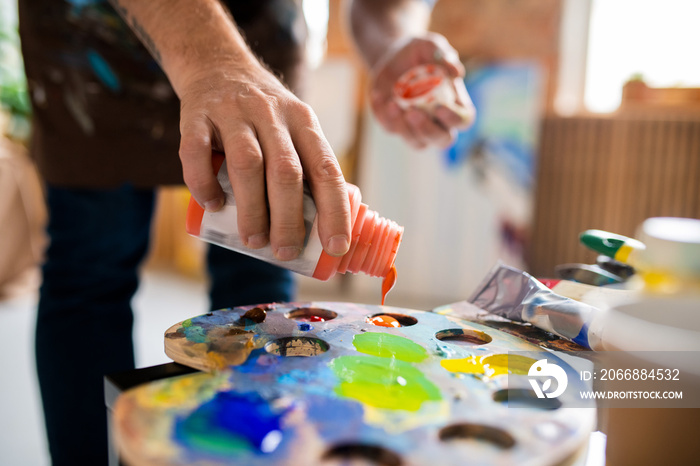 Young male painter adding gouache of orange color in palette before mixing