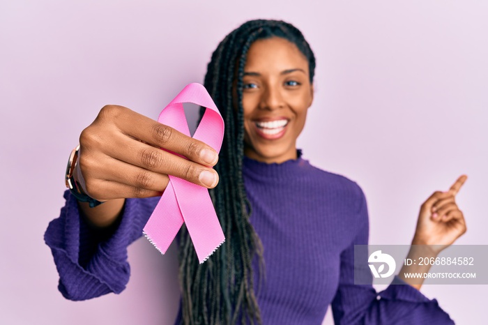 African american woman holding pink cancer ribbon smiling happy pointing with hand and finger to the side