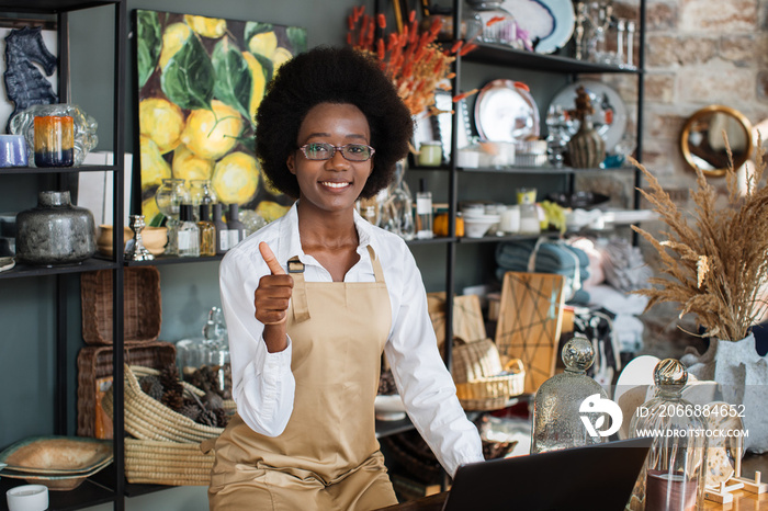 Beautiful afro american woman showing thumb up and smiling on camera while working at decor shop with laptop. Concept of people, technology and sale.