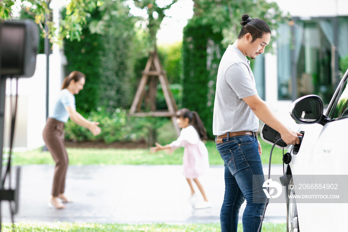 Focus image of progressive man charging electric car from home charging station with blur mother and daughter playing together in the background.