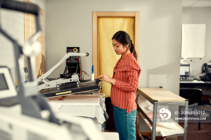 Printing expert. Young cheerful female worker using heat press transfer machine for printing advertisement sticker with text on t-shirt at workplace