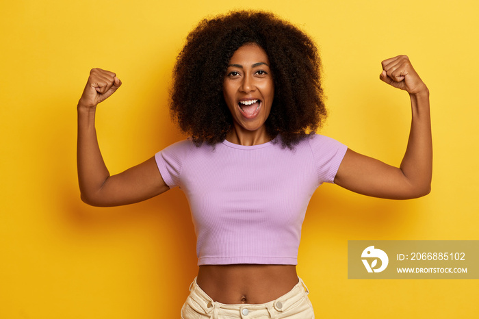 Overjoyed smiling woman with hands raised up in air, celebrates her achievement, dressed in casual purple T-shirt and jeans, isolated over yellow background