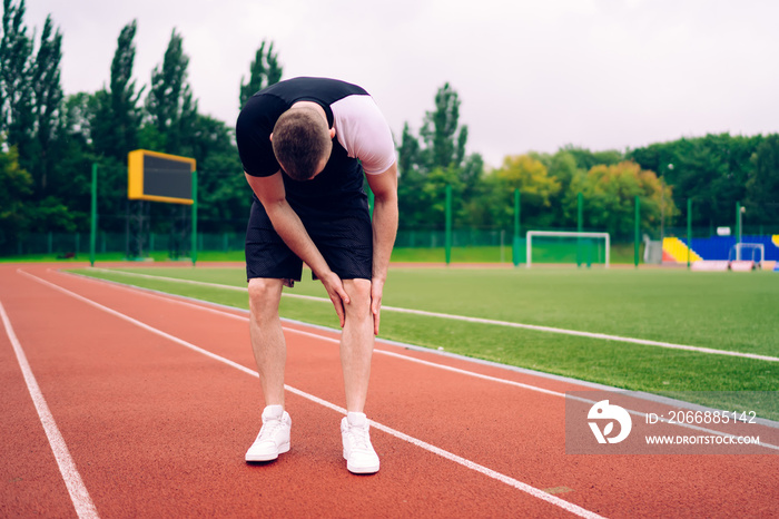 Sad caucasian male athlete having pain in knee after training doing massage for reducing ache, upset man runner in activewear having trouble with muckles after injury standing on stadium feel hurt