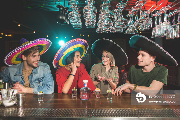 Happy girls and cheerful males drinking alcohol liquid. They wearing sombreros. Theme party in club concept
