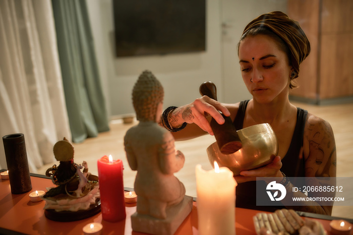 Serious young woman playing singing bowl beside altar