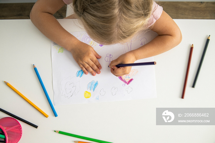 Blonde child girl draws with colored pencils sitting at the table. Top view, flat lay