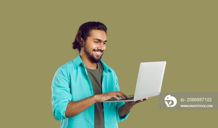 Studio shot of happy Indian man in casual turquoise shirt working on laptop computer. Cheerful handsome young ethnic IT professional, businessman, entrepreneur or web designer using modern notebook PC