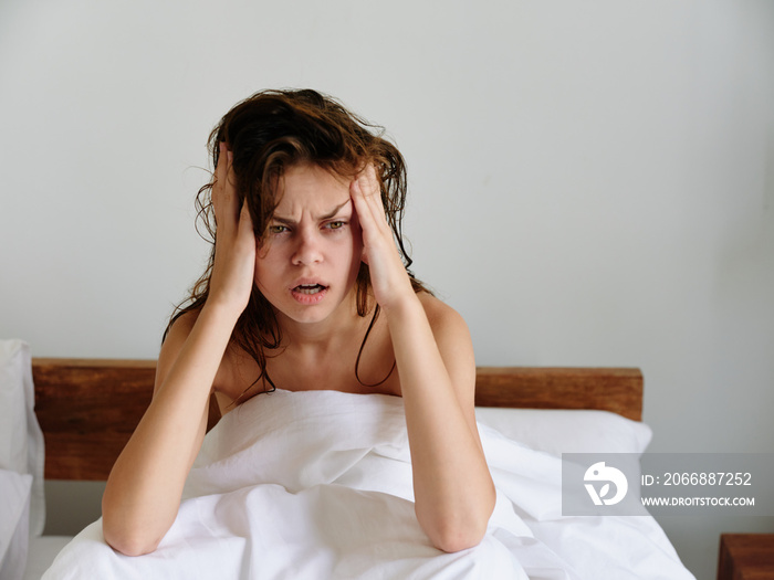 A woman sits on a bed in the bedroom and covers her body with a blanket, early awakening, sleepy appearance, rubbing her eyes from sleep, fatigue and depression